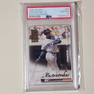 May include: A 1998 Studio Masterstrokes baseball card featuring Ken Griffey Jr. in a Seattle Mariners uniform. The card is graded GEM MT 10 and has a PSA grade of 56045521.