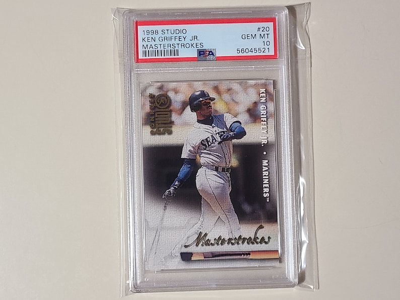 May include: A 1998 Studio Masterstrokes baseball card featuring Ken Griffey Jr. in a Seattle Mariners uniform. The card is graded GEM MT 10 and has a PSA grade of 56045521.