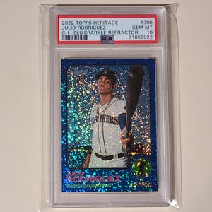 May include: A baseball card featuring Julio Rodriguez in a Mariners uniform, holding a bat. The card has a blue sparkle refractor design and is encased in a protective holder. The card's details include the 2022 Topps Heritage series and a GEM MT 10 grade.