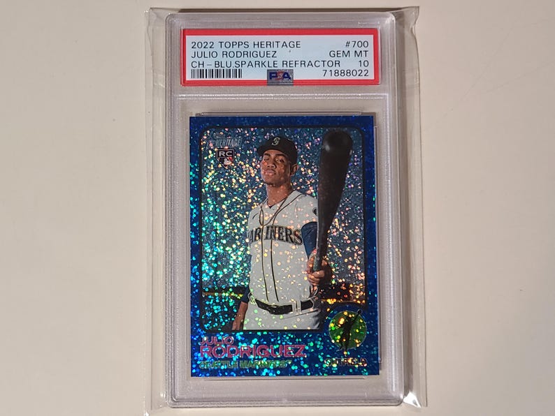 May include: A baseball card featuring Julio Rodriguez in a Mariners uniform, holding a bat. The card has a blue sparkle refractor design and is encased in a protective holder. The card's details include the 2022 Topps Heritage series and a GEM MT 10 grade.