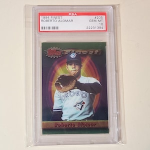 May include: A 1994 Topps Finest Roberto Alomar baseball card, graded PSA GEM MT 10. The card features a portrait of the player in a Toronto uniform with a glove, set against a dark green and purple background with the text Roberto Alomar.