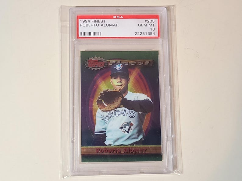 May include: A 1994 Topps Finest Roberto Alomar baseball card, graded PSA GEM MT 10. The card features a portrait of the player in a Toronto uniform with a glove, set against a dark green and purple background with the text Roberto Alomar.