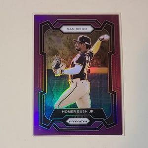 May include: A baseball card featuring Homer Bush Jr. in a throwing pose. The card has a purple and iridescent design with the text "SAN DIEGO" at the top and "HOMER BUSH JR." at the bottom. The card is from the Panini Prizm series.