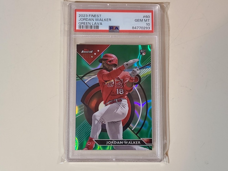 K&ouml;nnte beinhalten: Eine Baseball-Sammelkarte von Jordan Walker in einer Schutzh&uuml;lle. Die Karte zeigt einen Spieler in roter Uniform mit der Nummer 18, vor einem gr&uuml;nen und roten Hintergrund. Die Karte ist mit "2023 Finest" und "Green Lava" beschriftet.