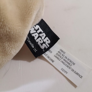 May include: Close-up of a plush, beige fabric with a black Star Wars tag. The tag includes the Star Wars logo, "Lucasfilm Ltd.", and product information, including "Surface Washable".