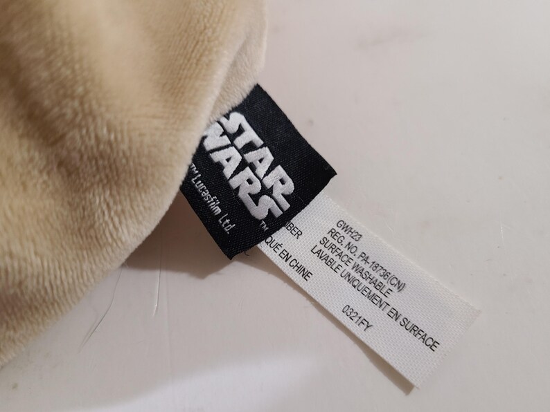 May include: Close-up of a plush, beige fabric with a black Star Wars tag. The tag includes the Star Wars logo, "Lucasfilm Ltd.", and product information, including "Surface Washable".