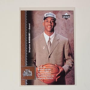 May include: A basketball trading card featuring Stephon Marbury in a suit, holding a Spalding NBA basketball. The card has text that reads "Stephon Marbury Guard" and "DECK". The card is in a protective sleeve.