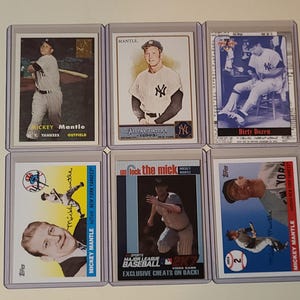 May include: Six collectible Mickey Mantle baseball cards encased in clear protective plastic sleeves. The cards feature various photographic and illustrated portraits of the player in his New York Yankees uniform, along with text including names and team details.