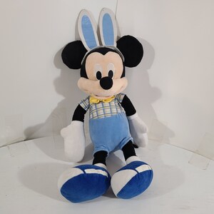 May include: A plush Mickey Mouse toy wearing blue bunny ears, a yellow bow tie, and a blue and yellow plaid shirt. The toy is sitting with its legs crossed and has blue shoes.