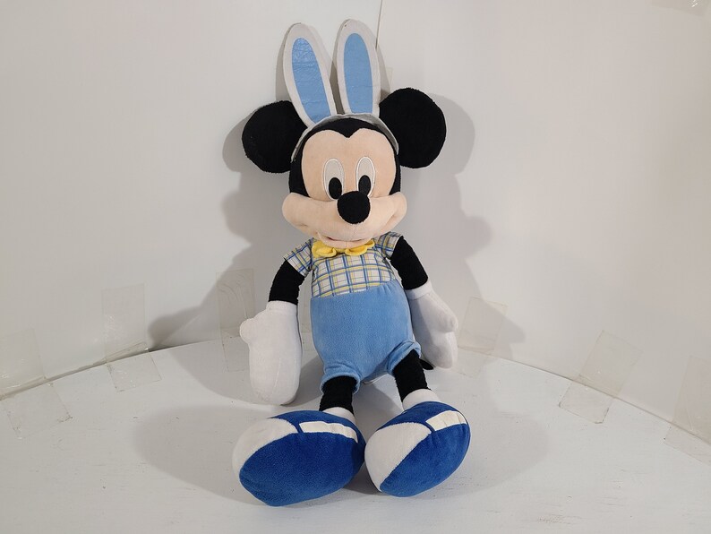 May include: A plush Mickey Mouse toy wearing blue bunny ears, a yellow bow tie, and a blue and yellow plaid shirt. The toy is sitting with its legs crossed and has blue shoes.