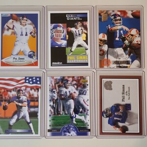 May include: Six different trading cards featuring Phil Simms, a former quarterback for the New York Giants. The cards are in protective plastic cases. The cards feature different photos of Simms in his Giants uniform, with the team logo and the words "New York Giants" and "Phil Simms" on each card.
