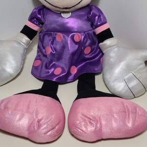 Pu&ograve; includere: Un giocattolo di peluche a forma di topo dei cartoni animati che indossa un vestito viola con pois rosa e guanti argentati. Il topo ha scarpe rosa scintillanti.