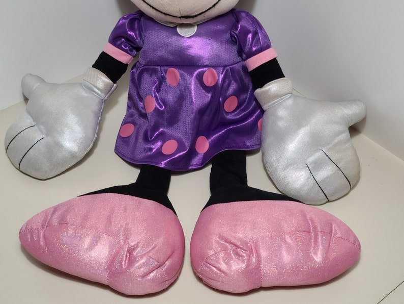 Pu&ograve; includere: Un giocattolo di peluche a forma di topo dei cartoni animati che indossa un vestito viola con pois rosa e guanti argentati. Il topo ha scarpe rosa scintillanti.