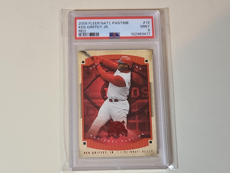 May include: A baseball trading card of Ken Griffey Jr. in a red and white Cincinnati Reds uniform, swinging a bat. The card is from the 2005 Fleer Nat'l Pastime series and is graded as Mint 9. The card is encased in a protective plastic sleeve.