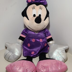 Pu&ograve; includere: Un grande giocattolo di peluche di Minnie Mouse che indossa un vestito viola con pois rosa e un fiocco rosa con pois bianchi. Ha guanti di glitter argentati e scarpe di glitter rosa.
