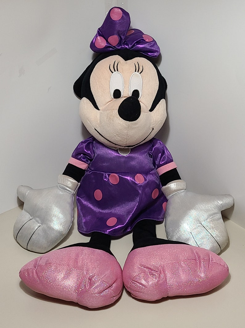 Pu&ograve; includere: Un grande giocattolo di peluche di Minnie Mouse che indossa un vestito viola con pois rosa e un fiocco rosa con pois bianchi. Ha guanti di glitter argentati e scarpe di glitter rosa.