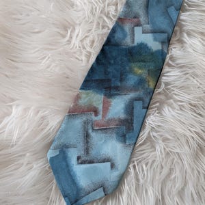 Peut inclure: Une cravate bleue avec un motif géométrique dans les tons de bleu, gris et marron. La cravate est faite d'un tissu lisse et est présentée sur une surface blanche et duveteuse.