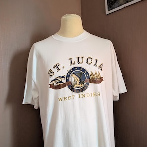 May include: White t-shirt featuring a blue and gold graphic of a marlin fish, dolphins, and a sailboat. The graphic is encircled by a blue circle with gold stars and the text "ST. LUCIA ISLAND PARADISE WEST INDIES".