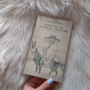 Puede incluir: Un libro vintage de Penguin Classics titulado "Antología de la literatura japonesa". La portada presenta una ilustración en blanco y negro de una figura vestida montando un burro. El libro está sobre un fondo blanco y esponjoso.