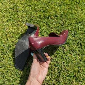 May include: A pair of burgundy leather heeled shoes with a black heel and a white stitch detail. The shoes are being held by a hand.