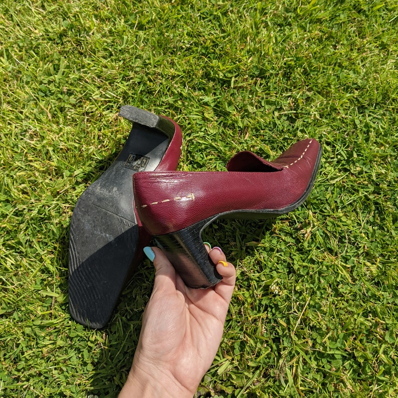May include: A pair of burgundy leather heeled shoes with a black heel and a white stitch detail. The shoes are being held by a hand.