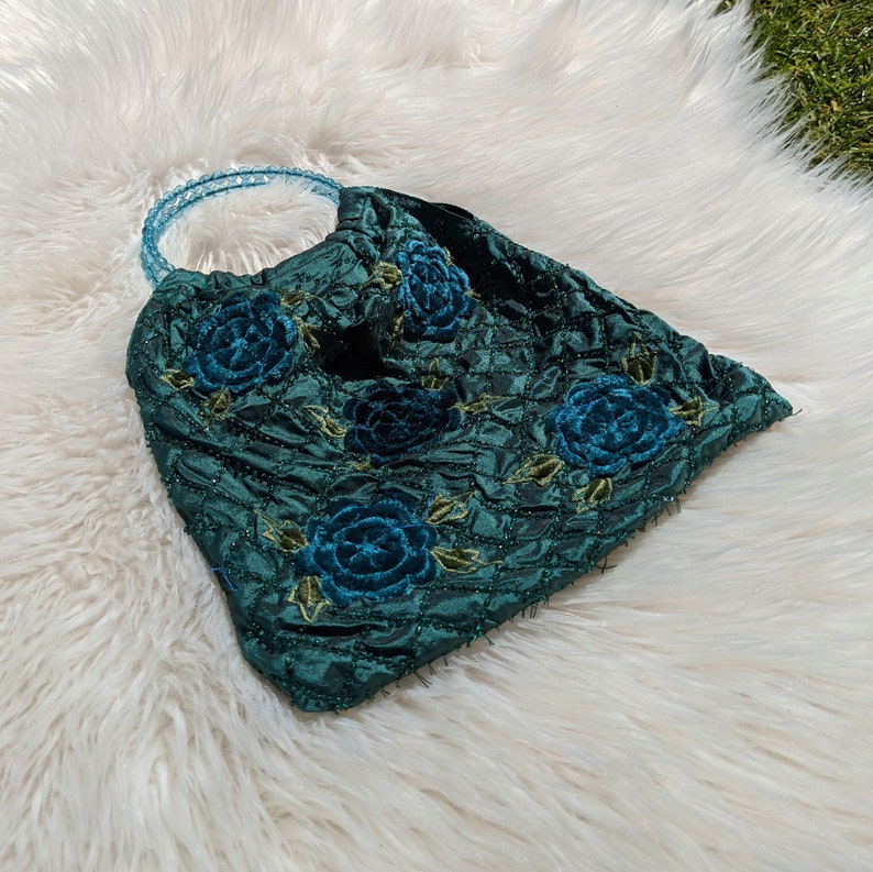 Pode incluir: Uma bolsa acolchoada azul-esverdeada com uma al&ccedil;a de pl&aacute;stico transparente e rosas de veludo azul bordadas.
