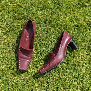 May include: Two pairs of burgundy leather shoes with white stitching. The shoes have a pointed toe and a low heel. The shoes are on a green grassy surface.