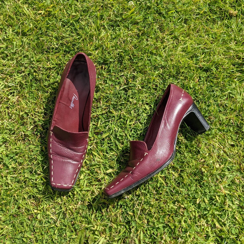 May include: Two pairs of burgundy leather shoes with white stitching. The shoes have a pointed toe and a low heel. The shoes are on a green grassy surface.