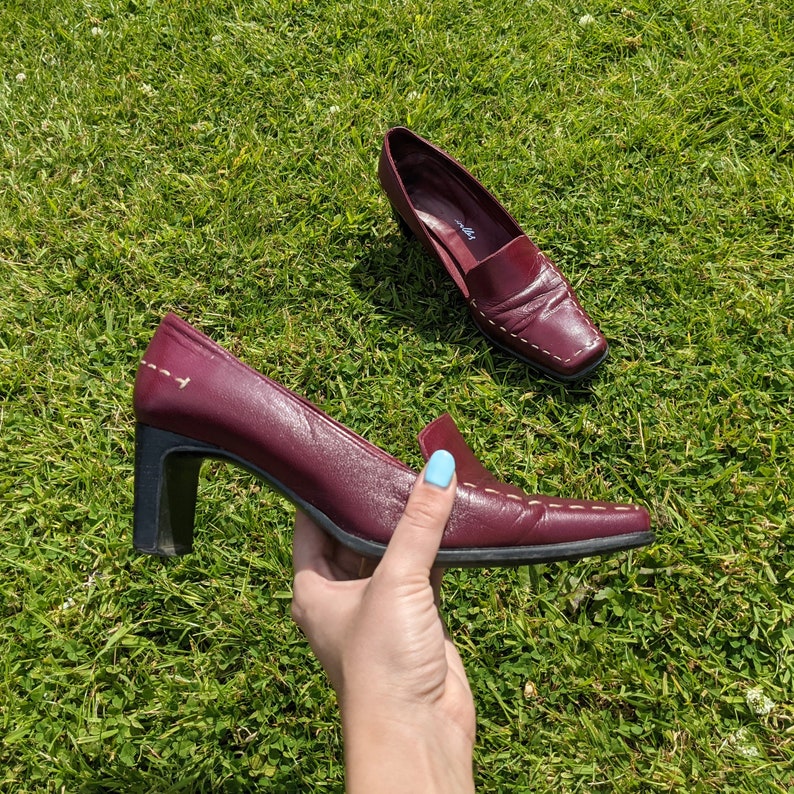 May include: A pair of burgundy leather heeled shoes with a pointed toe and a decorative stitch detail. The shoes have a low block heel.