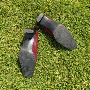 May include: A pair of burgundy leather heeled shoes with black soles. The shoes are on a green grassy surface.