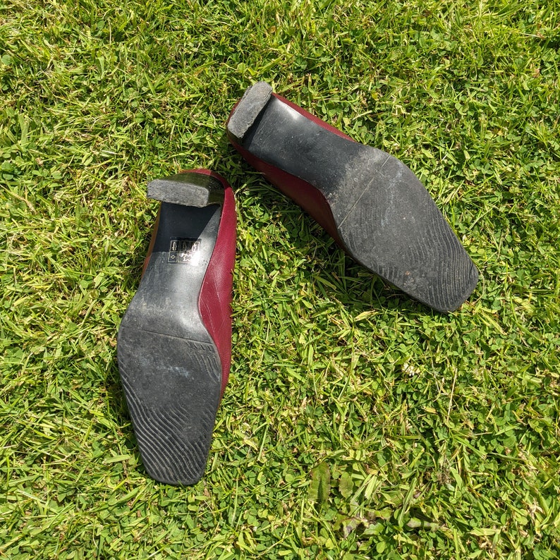 May include: A pair of burgundy leather heeled shoes with black soles. The shoes are on a green grassy surface.