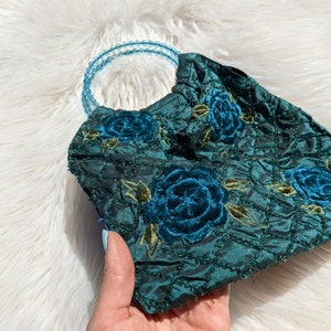 Pode incluir: Uma bolsa de m&atilde;o de veludo verde escuro com uma al&ccedil;a de contas azuis e bordado floral. As flores s&atilde;o em tons de azul e verde.