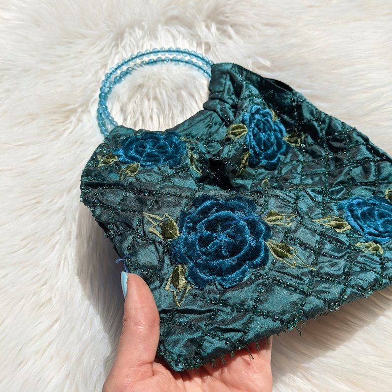 Pode incluir: Uma bolsa de m&atilde;o de veludo verde escuro com uma al&ccedil;a de contas azuis e bordado floral. As flores s&atilde;o em tons de azul e verde.