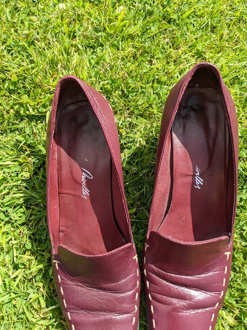 May include: A pair of burgundy leather loafers with white stitching. The loafers have a pointed toe and a low heel. The inside of the loafers is labeled "Cavalli".