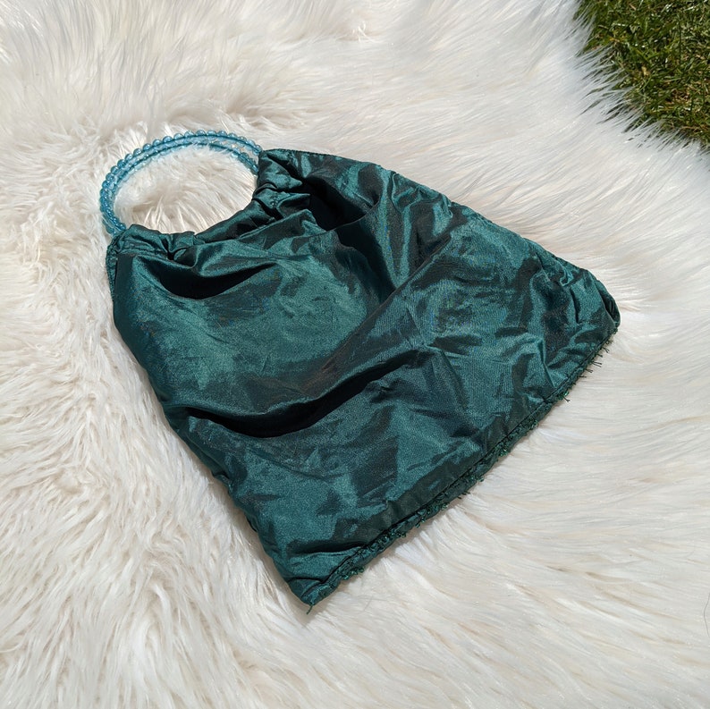 Pode incluir: Uma bolsa de tecido verde escuro com uma al&ccedil;a de contas azul claro. A bolsa &eacute; feita de um material macio e sedoso e tem um visual drapeado e plissado.