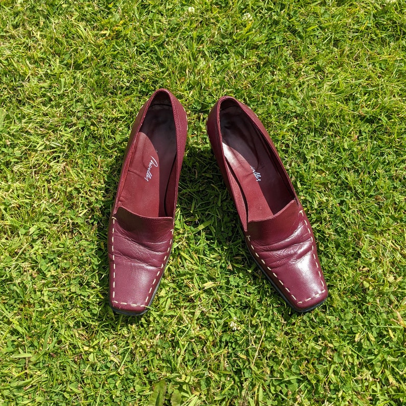 May include: A pair of burgundy leather loafers with white stitching. The loafers have a square toe and a low heel. The brand name "Joella" is visible on the inside of the left shoe.