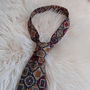 Op de afbeelding: Een stropdas met patroon en een knoop. De stropdas heeft een herhalend geometrisch ontwerp in tinten blauw, bordeauxrood en goud. De stropdas is gemaakt van een zijdeachtig materiaal en wordt gepresenteerd op een witte, zachte ondergrond.