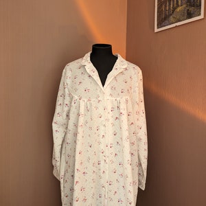 Peut inclure: Chemise de nuit blanche à manches longues ornée d'un motif floral de petites fleurs rouges et roses. La chemise de nuit a un devant boutonné, un col et est présentée sur un mannequin noir. Le tissu semble doux et léger.