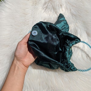 Pode incluir: Uma bolsa de m&atilde;o de cetim verde escuro com uma al&ccedil;a de contas. A bolsa tem um fecho redondo de cor prateada.