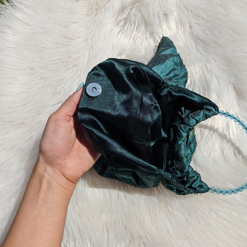 Pode incluir: Uma bolsa de m&atilde;o de cetim verde escuro com uma al&ccedil;a de contas. A bolsa tem um fecho redondo de cor prateada.