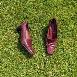 May include: A pair of burgundy leather heeled shoes with white stitching. The shoes have a pointed toe and a low heel.