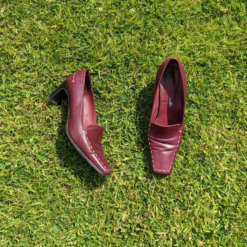 May include: A pair of burgundy leather heeled shoes with white stitching. The shoes have a pointed toe and a low heel.