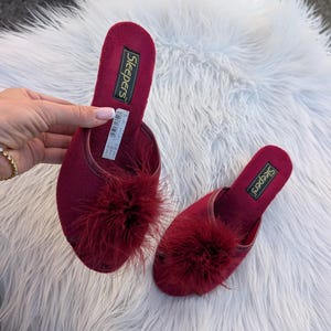 May include: Burgundy slippers with a fluffy feather detail. The slippers have the word "Sleepers" printed on the insole. The slippers are displayed on a white, fluffy surface.