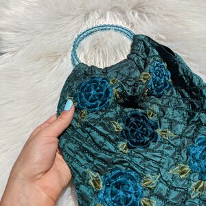 Pode incluir: Uma bolsa de m&atilde;o de veludo azul-esverdeado com uma al&ccedil;a de contas e bordado floral. As flores s&atilde;o de um azul-esverdeado escuro e t&ecirc;m um efeito 3D.