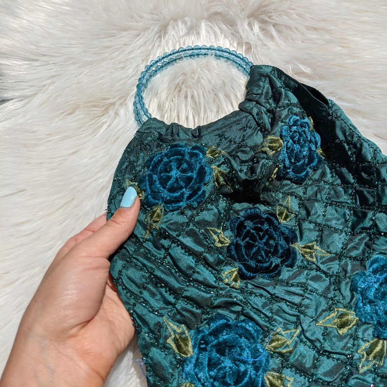 Pode incluir: Uma bolsa de m&atilde;o de veludo azul-esverdeado com uma al&ccedil;a de contas e bordado floral. As flores s&atilde;o de um azul-esverdeado escuro e t&ecirc;m um efeito 3D.