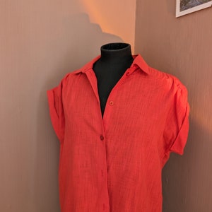 May include: A bright red button-down shirt with short sleeves, displayed on a black mannequin. The shirt has a classic collar and a textured fabric. The mannequin is positioned in a corner against a neutral-toned wall.