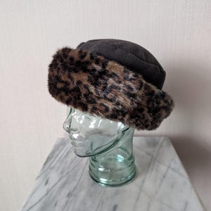 Puede incluir: Un sombrero gris oscuro con un ribete de piel sintética de leopardo. El sombrero se exhibe en una forma de cabeza de vidrio, mostrando su forma y diseño. El ribete de piel del sombrero presenta un patrón de manchas marrones y negras.