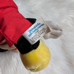 Może przedstawiać: Zabytkowa pluszowa zabawka Myszki Miki, z czerwoną kurtką, czarnymi spodenkami, ż&oacute;łtymi butami i białymi rękawiczkami. Etykieta głosi "Disneyland Walt Disney World" i "Disney". Pluszak leży na białej, puszystej powierzchni.