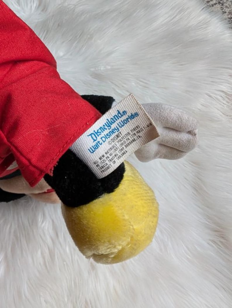 Może przedstawiać: Zabytkowa pluszowa zabawka Myszki Miki, z czerwoną kurtką, czarnymi spodenkami, ż&oacute;łtymi butami i białymi rękawiczkami. Etykieta głosi "Disneyland Walt Disney World" i "Disney". Pluszak leży na białej, puszystej powierzchni.