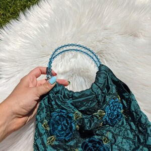 Pode incluir: Uma bolsa de m&atilde;o de veludo azul-esverdeado com bordados florais. A bolsa tem uma al&ccedil;a de contas feita de contas de vidro azul claro.